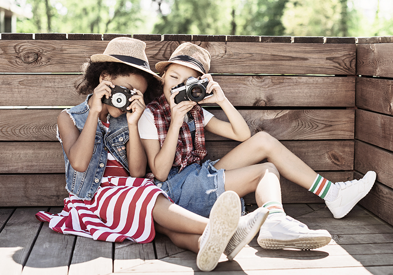 Två unga tjejer sitter bredvid varandra utomhus. Båda håller en kamera var som är framför ansiktet.